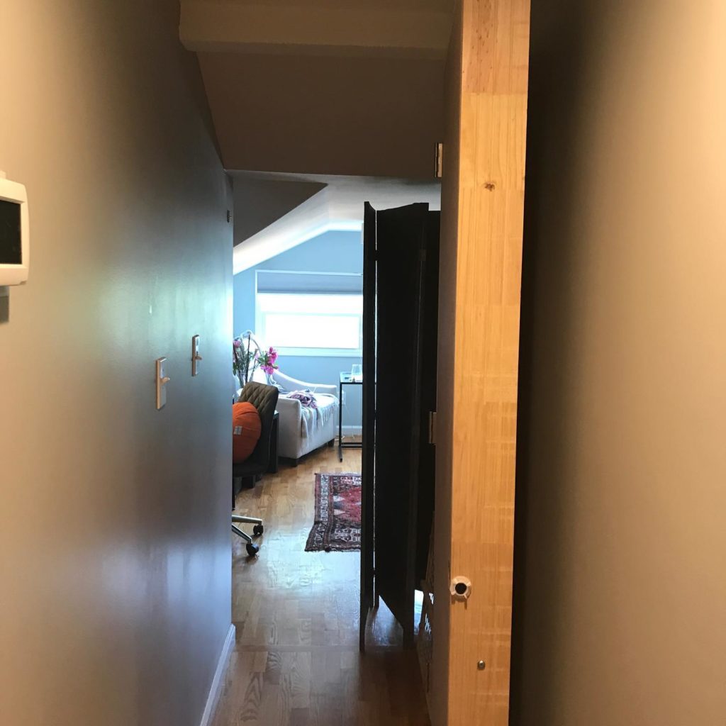 Hallway with an open door leading to a bright room with hardwood floors, a rug, and a seating area near a window.