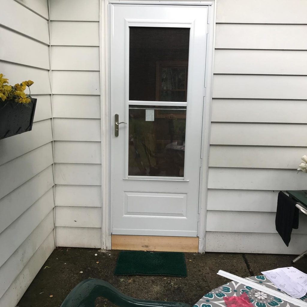 White storm door with a glass panel, set in white siding, with a small green doormat and patio furniture nearby.