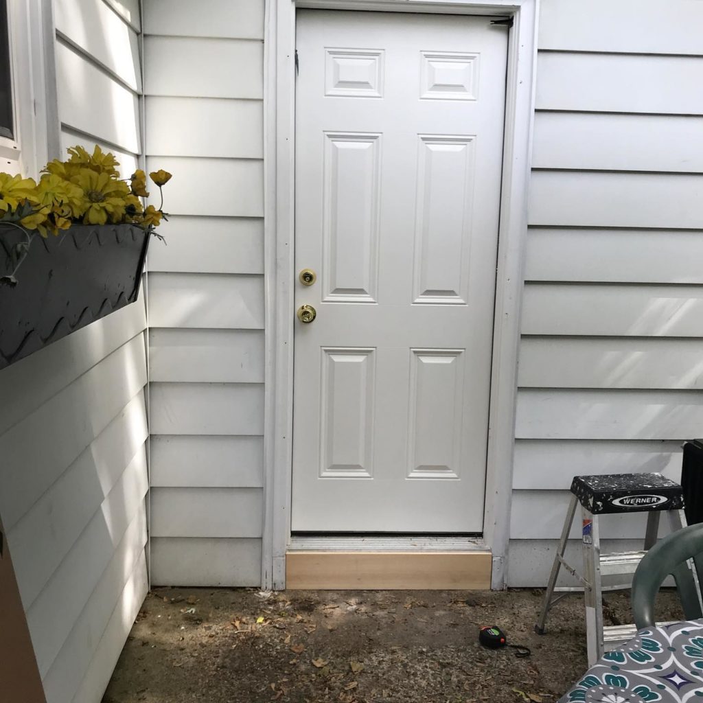 White exterior door surrounded by white siding, with a flower box on the left and a step stool on the right.